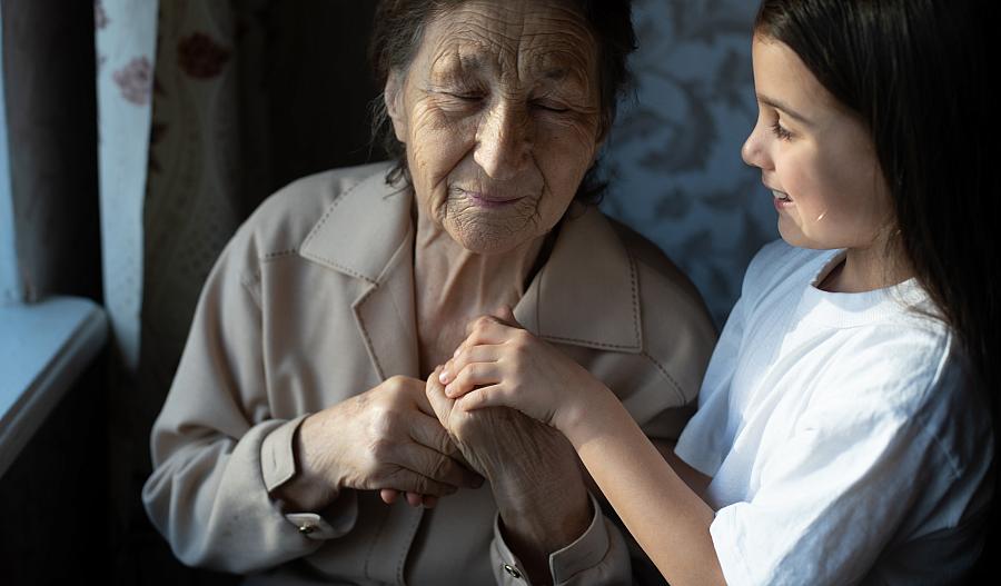 Una fotografía muestra a una mujer adulta mayor y a una niña tomadas de las manos. La niña está sonriendo mientras mira al rostro de la mujer, que tiene sus ojos cerrados. 